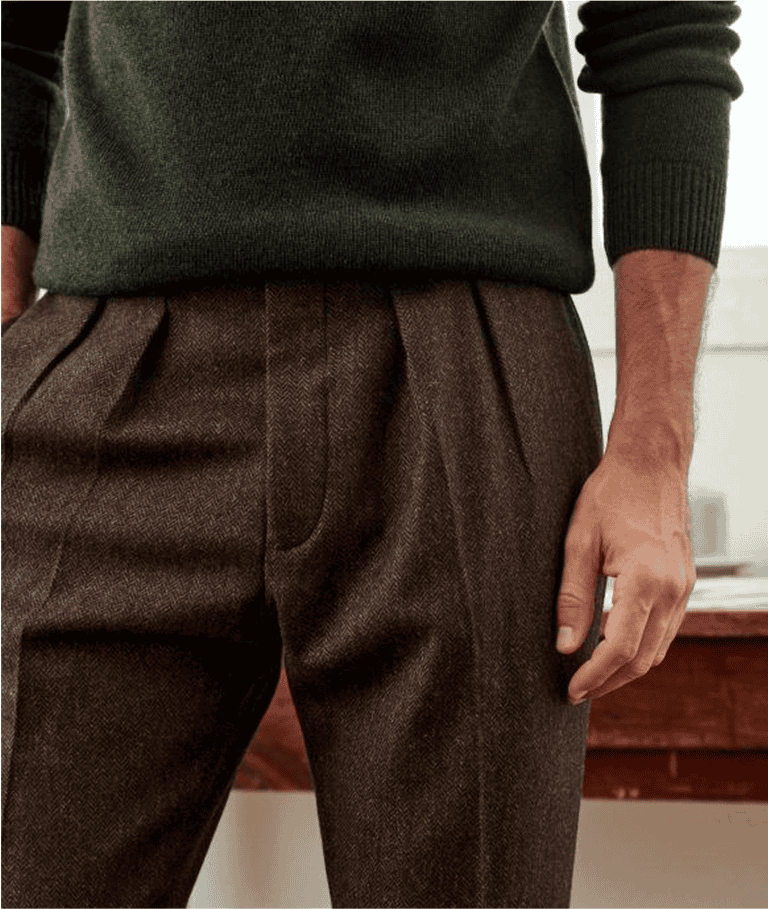 A man wearing brown pleated trousers and a dark green sweater stands indoors, partially shown from chest to thigh.